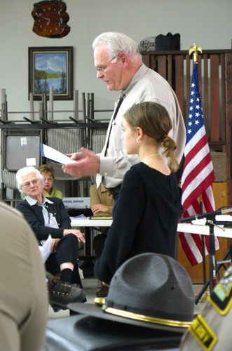 photo of a girl standing next to a man in a tie, he is reading the text of an award he is giving the girl. You can see attendees in the background and the shoulders of two sheriffs and a sheriff's hat in the foreground.
