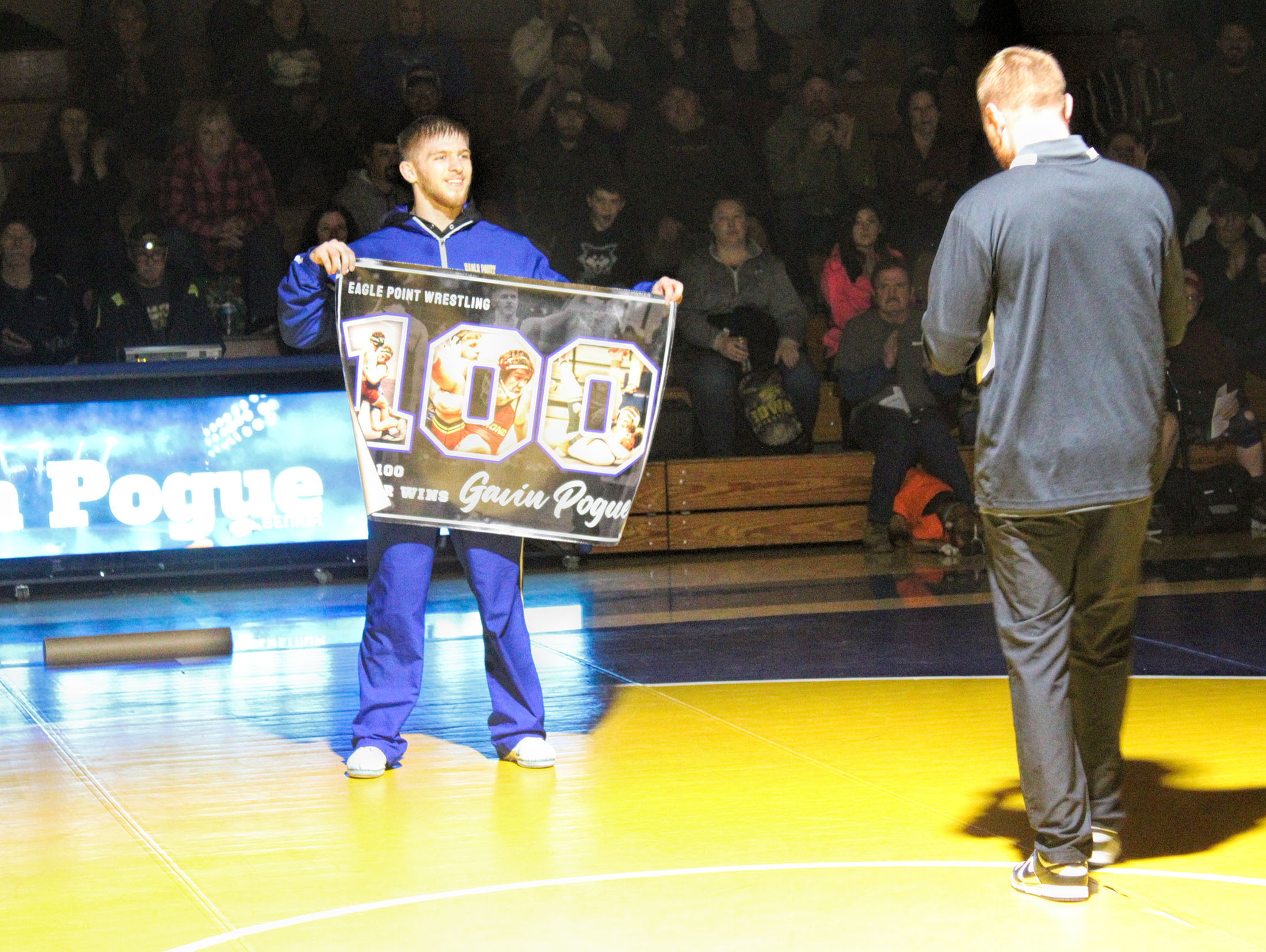 photo of a high school wearing a warm up suit. he is holding up a banner celebrating 100 matches.