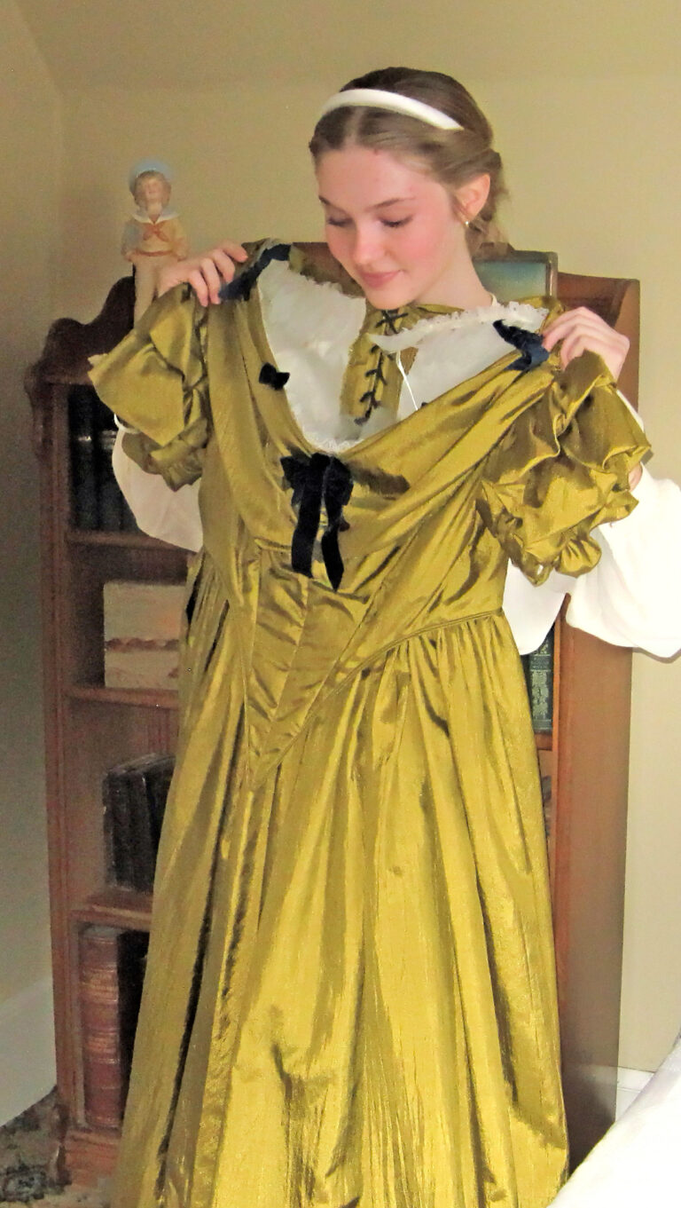 Photo of a young girl in historic costume holding a gold silken gown up in front of her as she looks in a mirror.