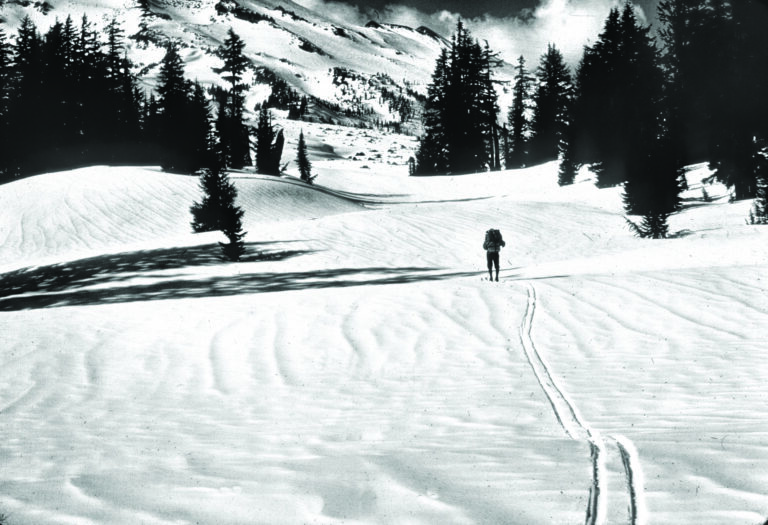 black and white photo of a man cross country skiing across a snowy meadow. there is a snow covered mountain in the background and conifer trees around.