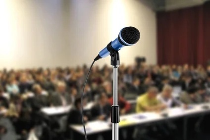 photo of a microphone on a stand with an audience in the background.