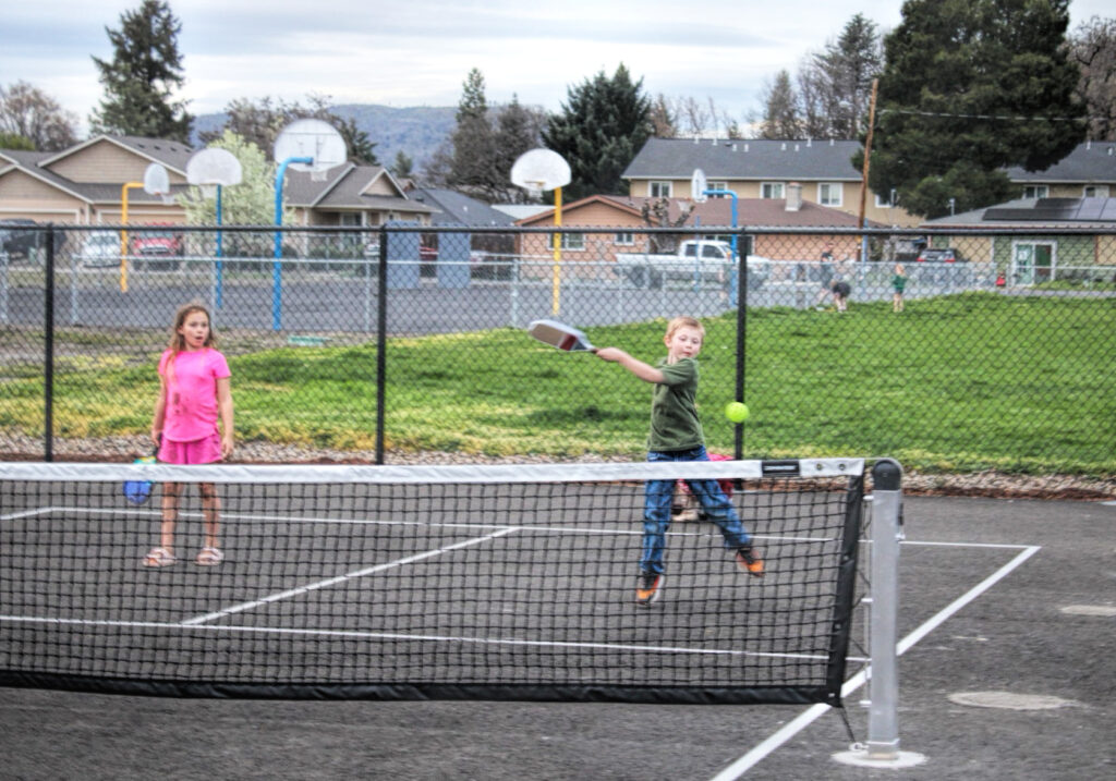 photo of two young children on a pickleball court. the boy has just hit the ball and it's headed over the net.