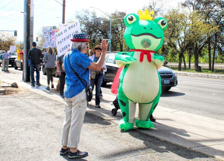photo of several no kings protestors holding signs and waiving at drivers. there is one protestor in an inflatable frog costume.