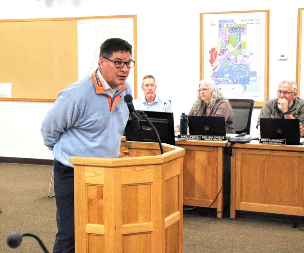 photo of a man speaking into a microphone at a podium with council members seated behind him.