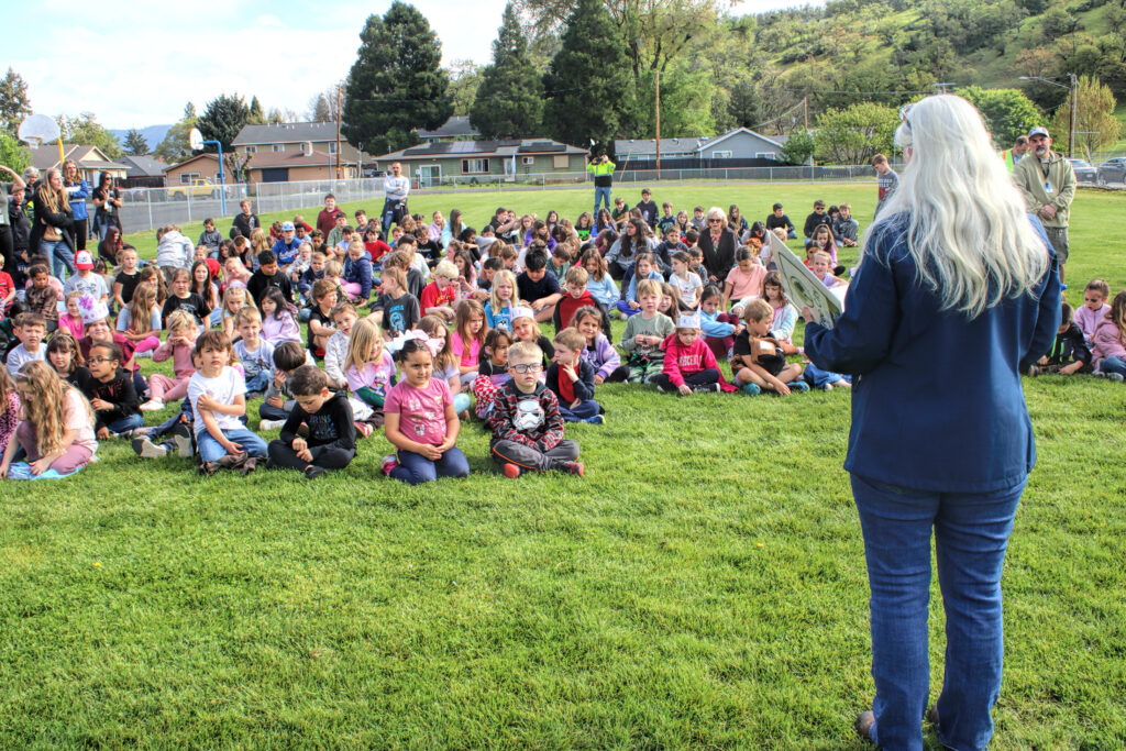 Photo of an adult woman standing in a grassy park in front of a large group of grade school children.