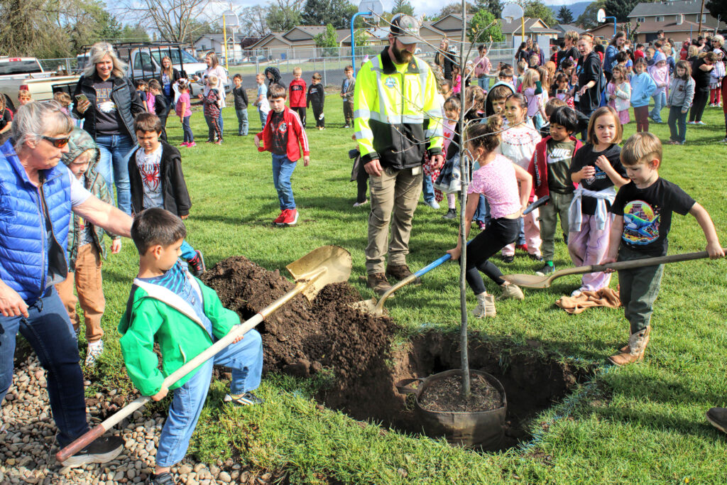 photo of a group of (supervised) children tossing dirt into a hole with a newly planted tree in it.