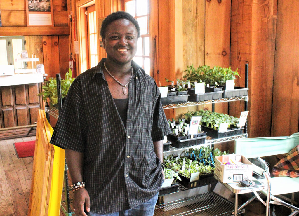 Photo of a man in front of a stand of small herbs for sale.