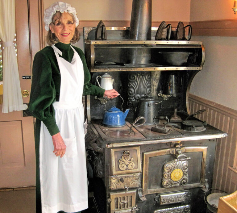 photo of a woman in period clothing standing next to a cast iron stove placing a teapot on a burner.
