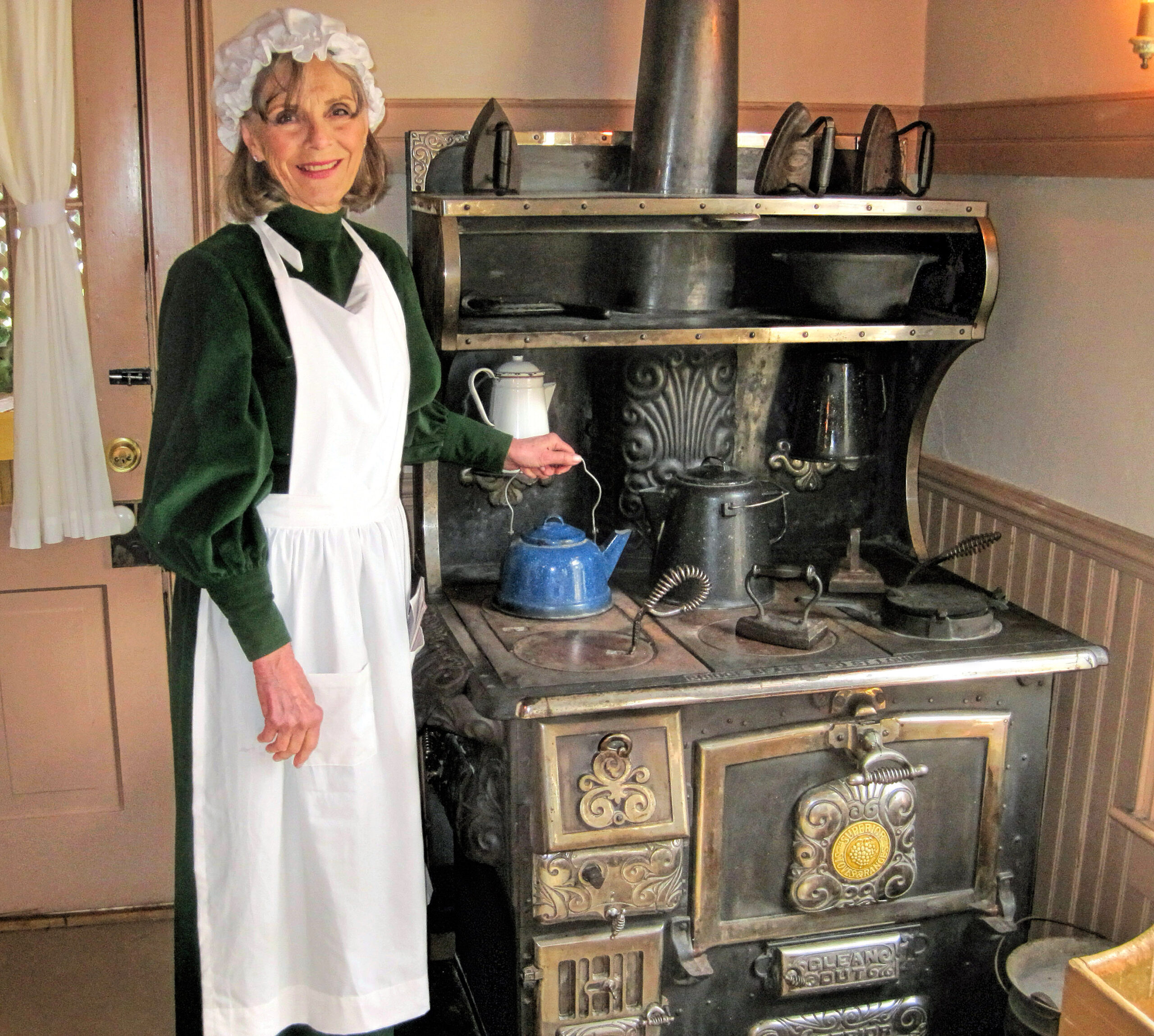 photo of a woman in period clothing standing next to a cast iron stove placing a teapot on a burner.