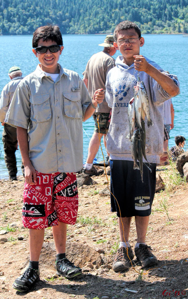 photo of two young boys in front of a lake, one is holding up a string of 8 or 9 freshly caught trout.