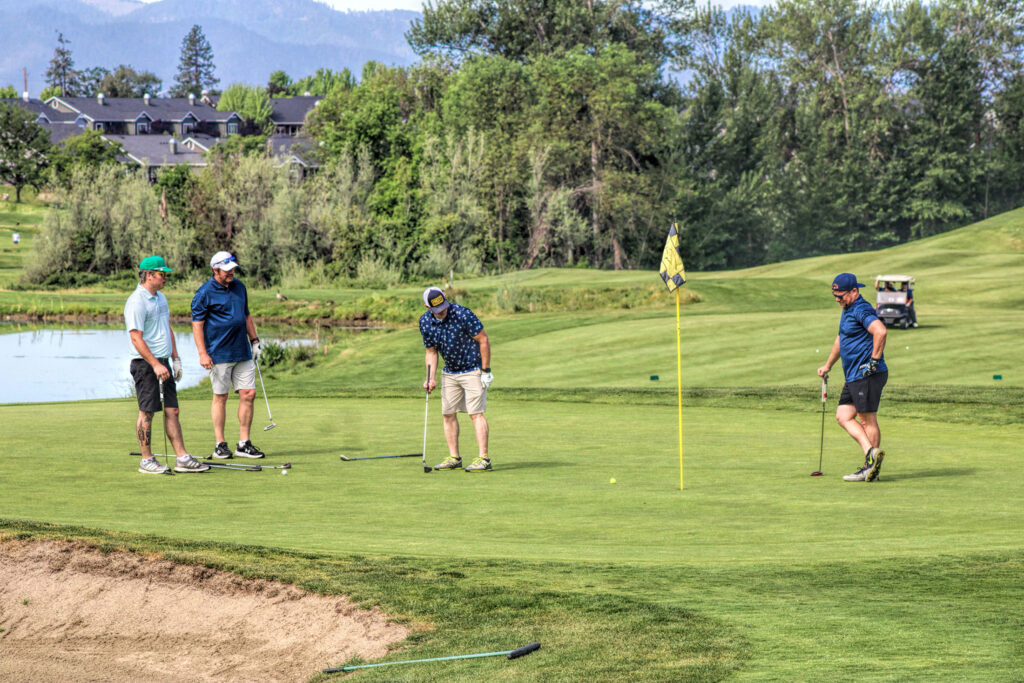 photo of four golfers on the putting green. one is putting, three are watching.
