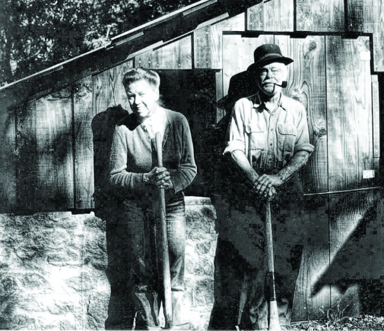 black and white photo of a couple in the late 60s standing in front of a wooden structure.