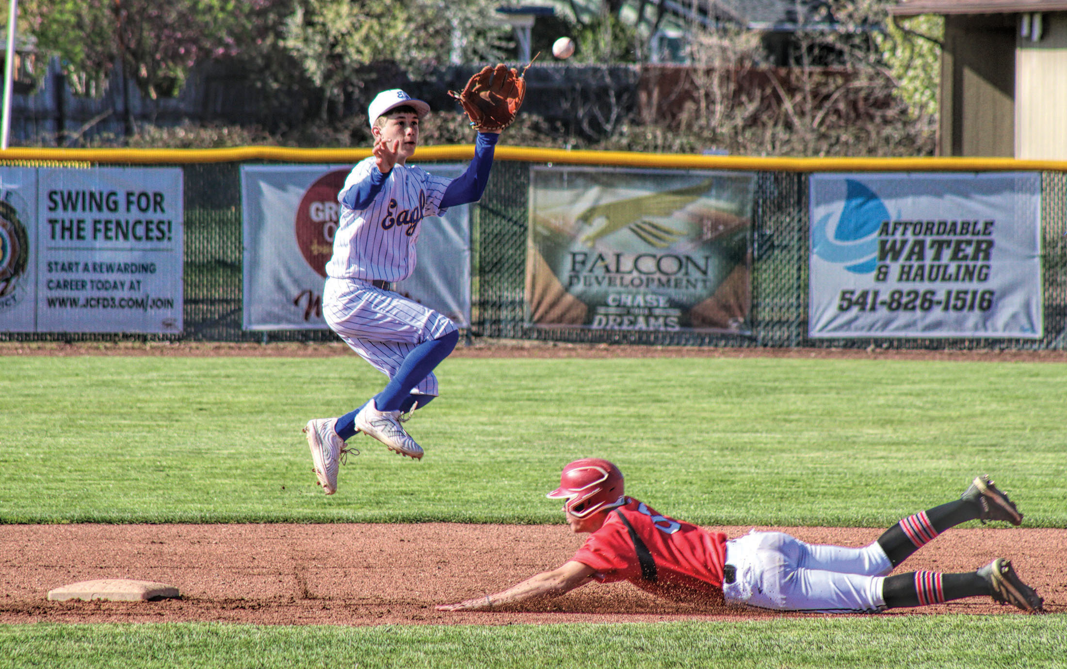 photo of a high school baseball game. one player is sliding into a base, the on base player is in mid air about to catch the ball.