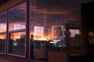 photo of the big glass window on a diner at sunset, the sunset reflects in the window and you can see a "for sale" sign.