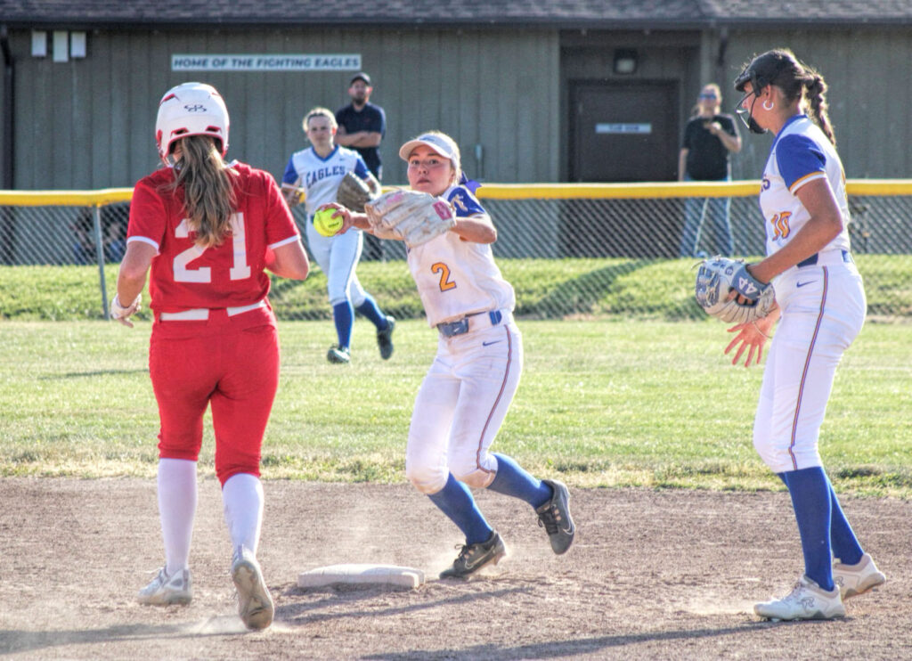 photo of a high school softball game. a player is winding back to throw the ball as the runner approaches the base.