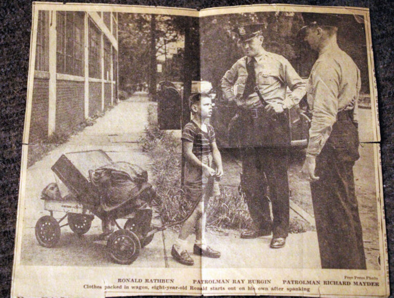 photo of an old newspaper clipping photo showing a young boy with belongings in a wagon looking up at one of two police officers talking to him.