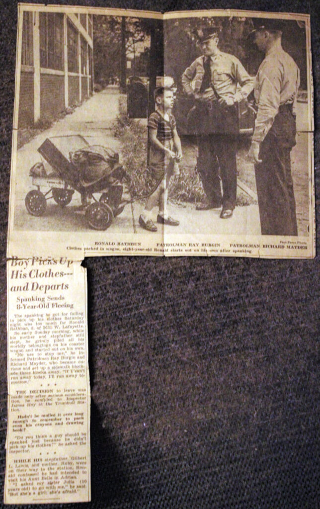 photo of an old newspaper clipping photo showing a young boy with belongings in a wagon looking up at one of two police officers talking to him. A short column accompanies the photo.