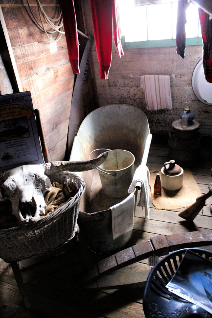 photo of the interior of the historic Woodhouse showing an old tin bathtub with a bucket inside and a steer skull in a basket at the foot of the tub.