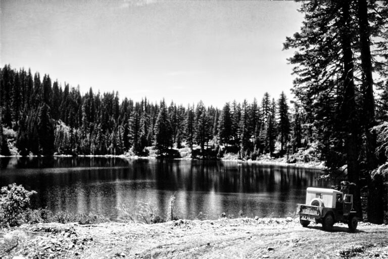 black and white photo of a small mountain lake surrounded by conifer trees. there is a small jeep parked near the shoreline.