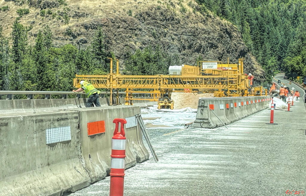 Photo of construction on a bridge. there is some large equipment and many workers in three-mile orange and three-mile yellow. there are mountains and conifer trees in the background.