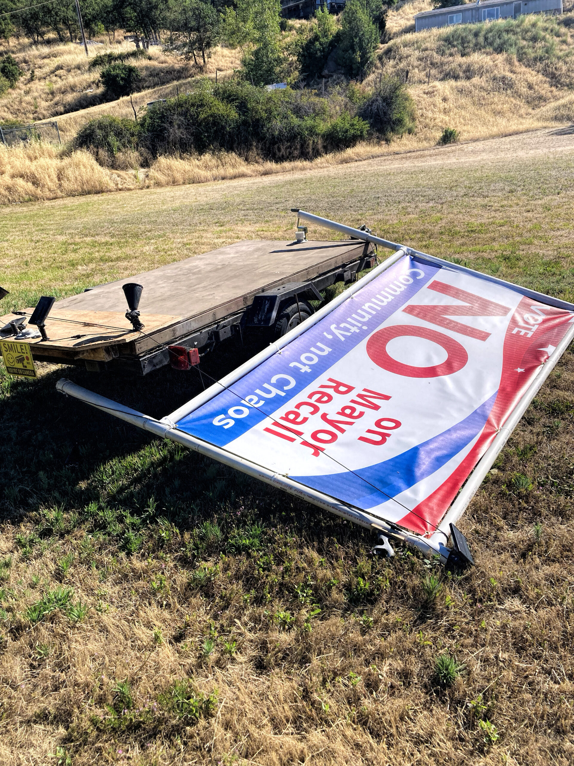 photo of a sturdy political sign ripped off of its wooden base and laying on the ground, wires connecting it to lighting hanging out. a smaller sign on the wooden platform reads "smile you're on camera"