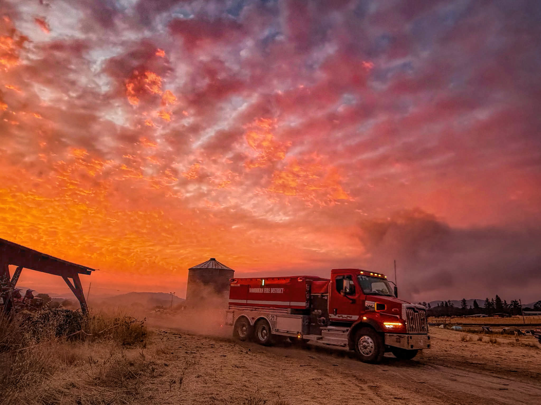 photo of a firetruck pulling out onto a dirt road with orange sky and a plume of smoke behind them.