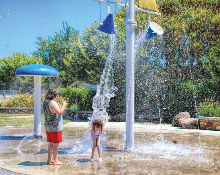 photo of one adult and one child at a small water park. a large bucket of water is splashing down on the child as sh ducks.
