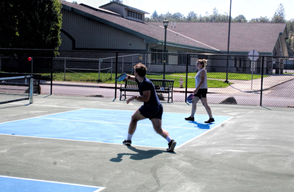 photo of a man and woman on a pickle ball court, the man has just hit the ball.