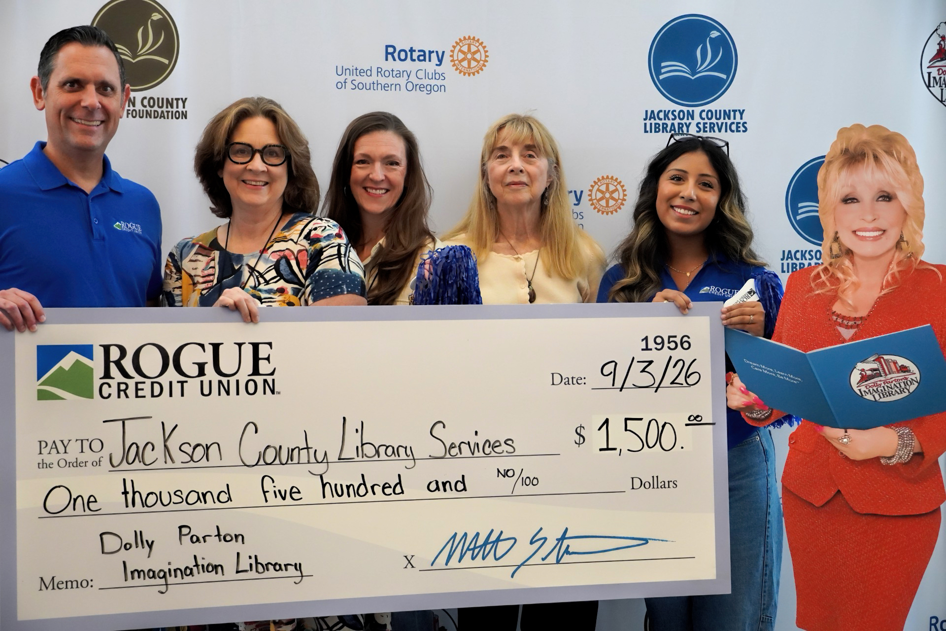 photo of five people holding a giant check in the amount of ,500 the memo reads :Dolly Parton Imagination Library. To their right is a life-sized cardboard Dolly