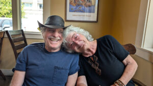 photo of a senior couple sitting in a cafe, the woman is leaning her head on the man's shoulder, both are laughing.