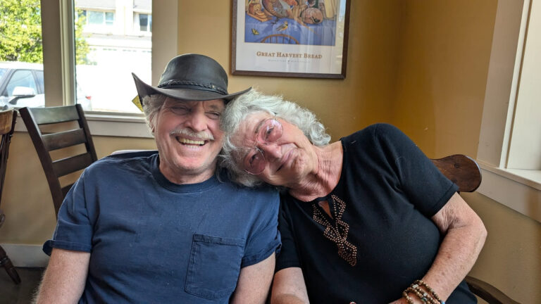 photo of a senior couple sitting in a cafe, the woman is leaning her head on the man's shoulder, both are laughing.