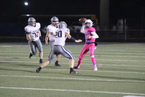photo of a high school football game, three players are running at a quarterback who has his arm up to throw the ball.
