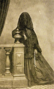 vintage black and white photo of a Victorian era female dressed in black with a black veil over her face. se is leaning on a banister.