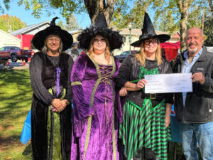 photo of three women dressed as witches and one man who is handing them an oversized check.