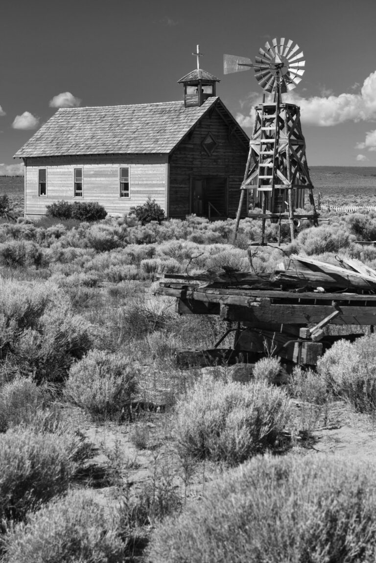 black and white photo of a church in a ghost town with a windmill and some old farming equipment.
