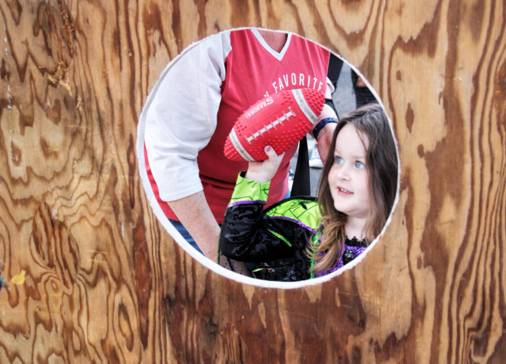 photo of a young girl in a spider witch costume peeking through a circle cut out of plywood. she's holding a football about to throw ith through the hole.