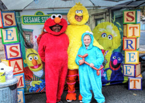 photo of two adults and one child dressed as sesame street characters Elmo, Big Bird and Cookie Monster. they are standing in a Sesame Street booth.